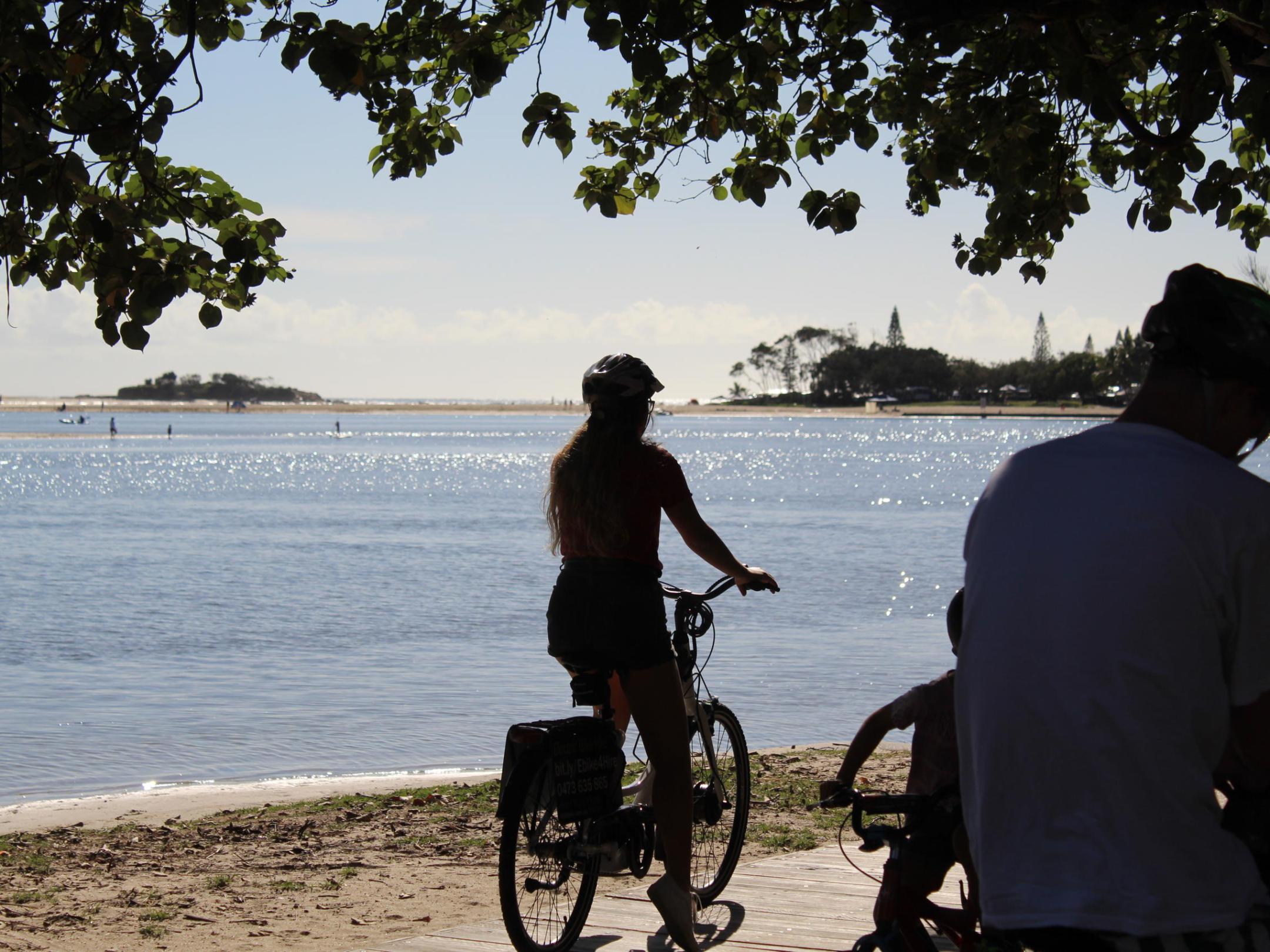 Couple enjoying a romantic weekend cycling EcoTekk hire e-bikes among cotton trees, with the Maroochy River mouth, Pincushion Island, and Cotton Tree Beach in the background.
