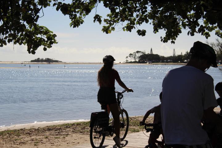 Couple enjoying a romantic weekend cycling EcoTekk hire e-bikes among cotton trees, with the Maroochy River mouth, Pincushion Island, and Cotton Tree Beach in the background.