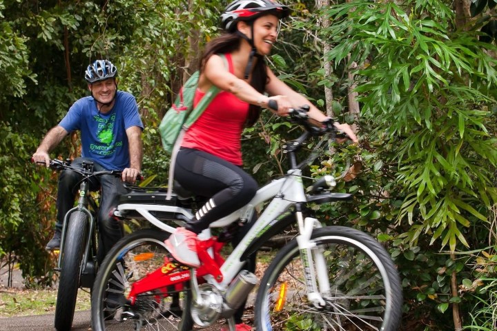 Couple enjoying a sunny day riding e-bikes on Noosa coastal pathway, laughing together.
