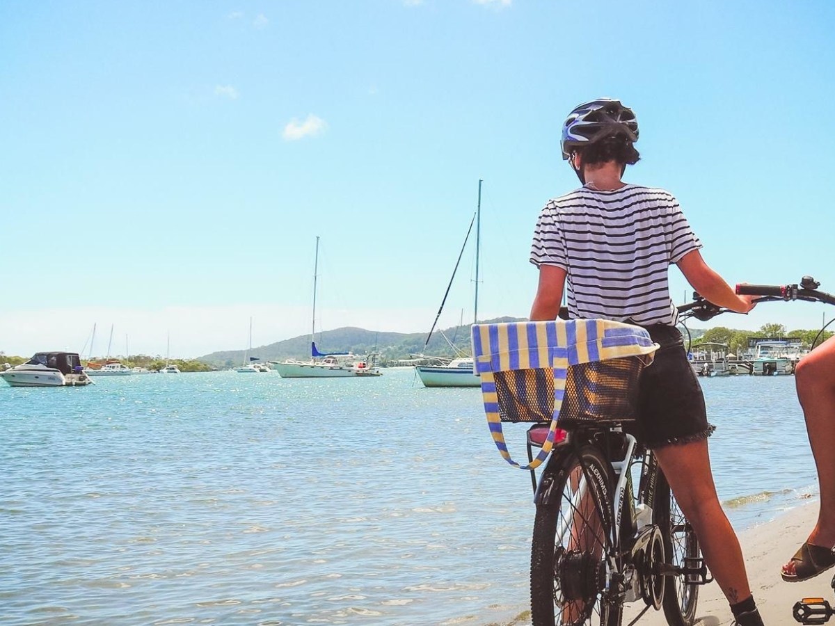 Two ladies on ecoTekk hired e-bikes enjoying the view across the turquoise waters of the Noosa River towards Pelican Beach Park and the Noosa River mouth, with boats and yachts moored and Noosa National Park hills in the background.