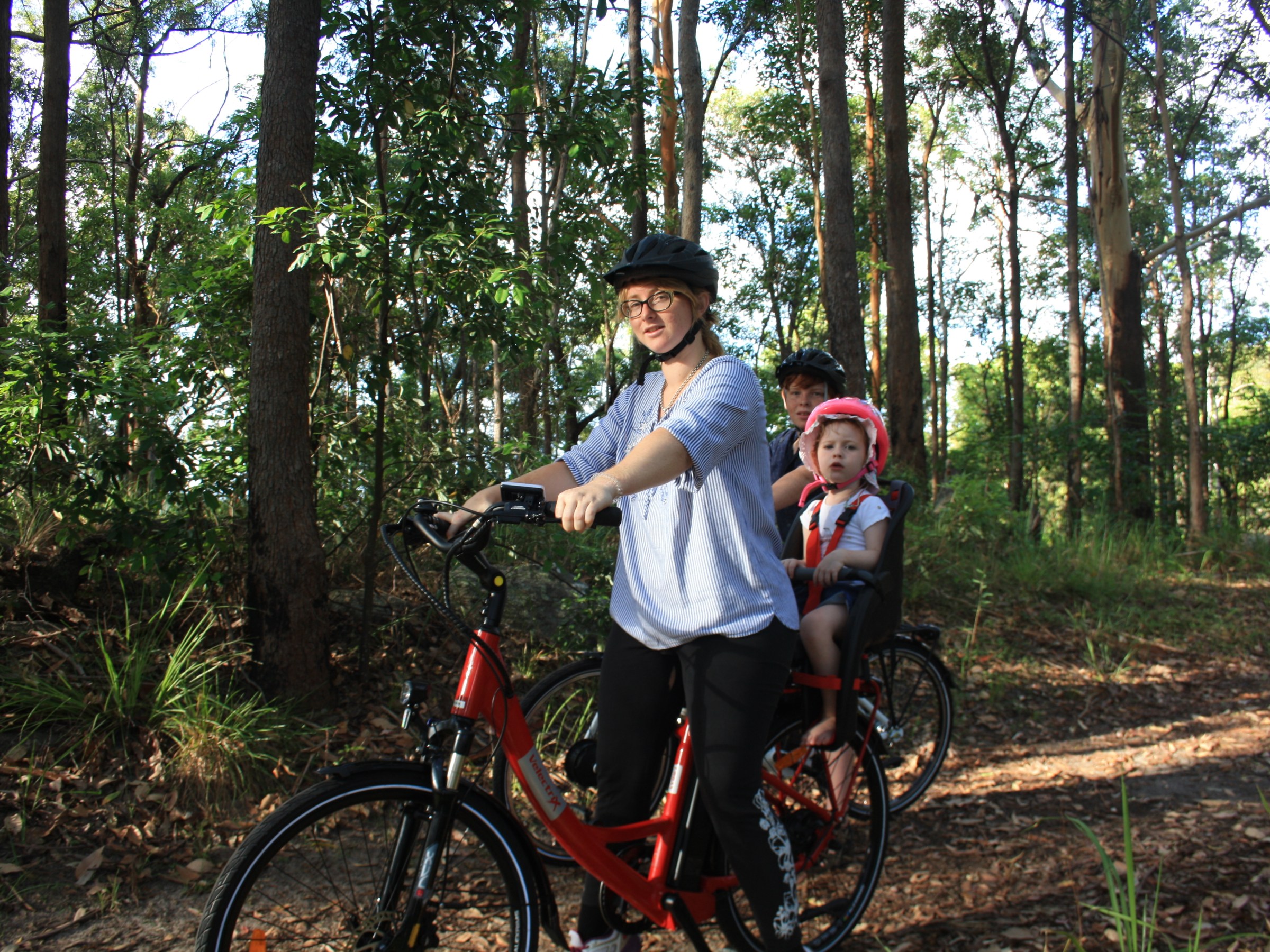 Mum with her daughter in a toddler bike seat and her uncle admire the Pink Bloodwood forest at Koala Park on EcoTekkSC e-bikes, with sunlight streaming through the trees.