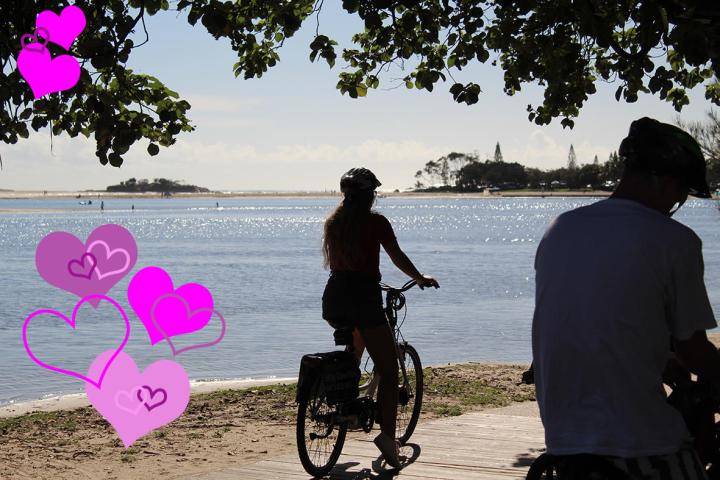 A couple riding e-bikes along the Maroochy River foreshore, with sparkling water in the background and love hearts overlaid, perfect for a Valentine's Day tour.