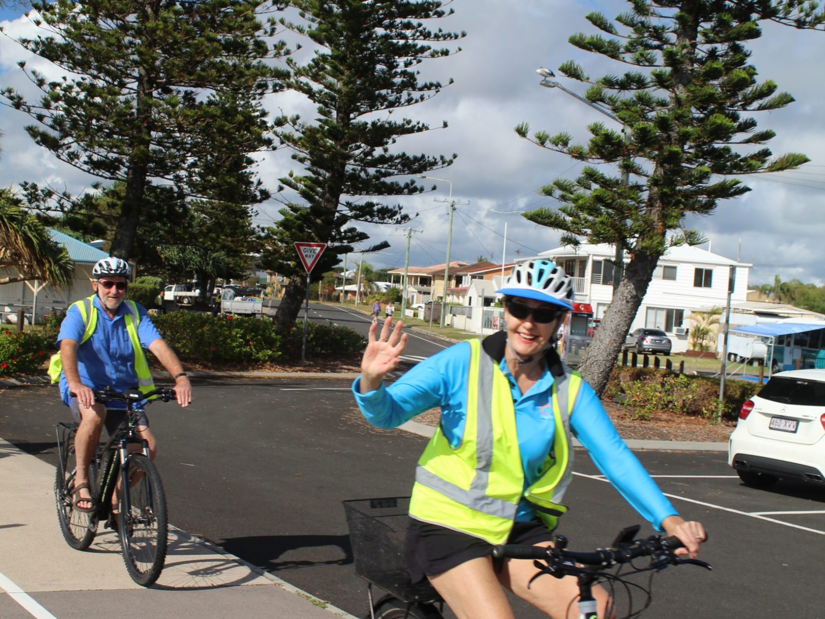 A group of mature riders on EcoTekkSC e-bikes, smiling and waving as they pass the Bunya Pine-lined section of the Coastal Pathway near Military Jetty, Golden Beach, Caloundra.