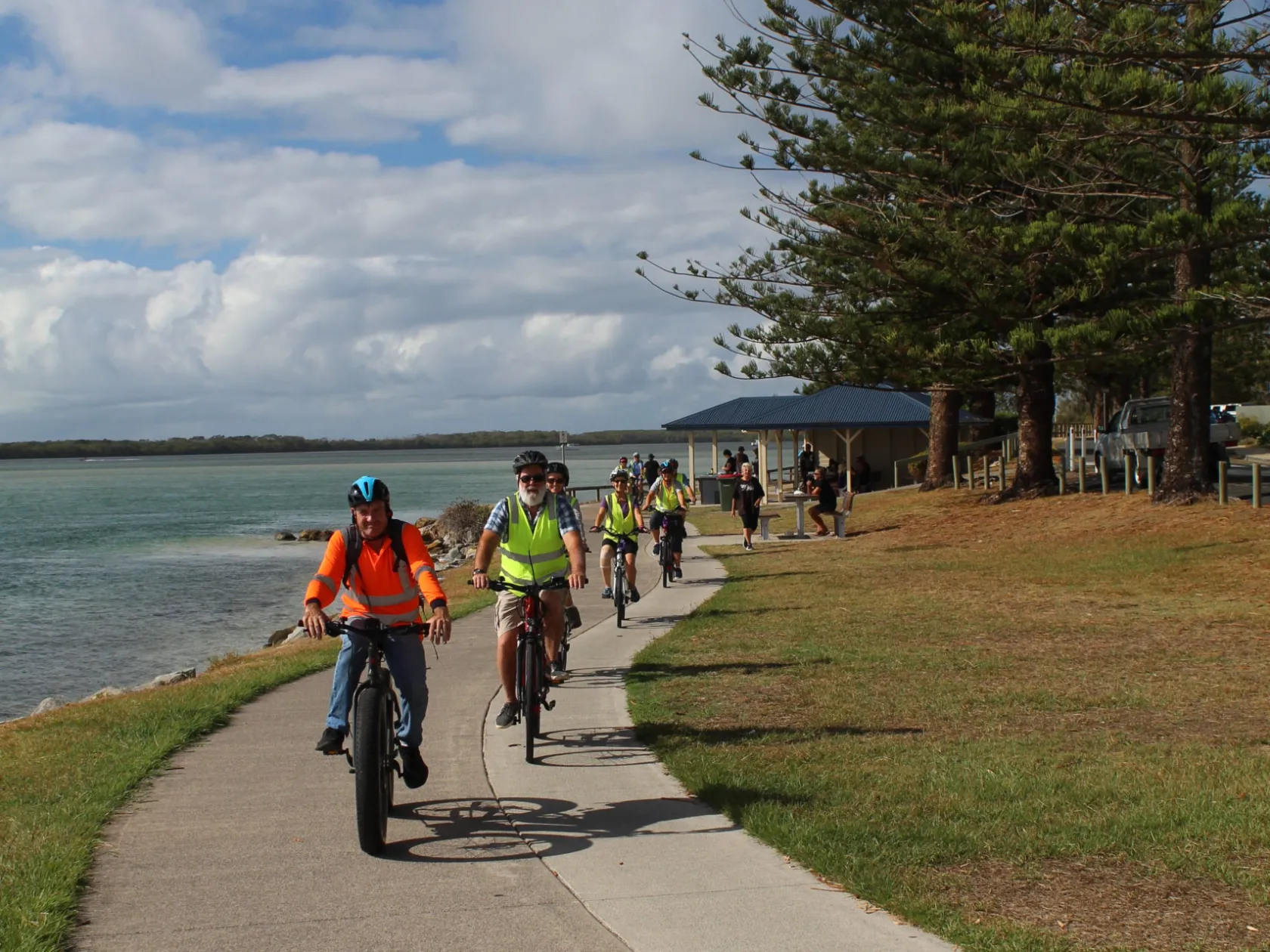 A group of mature riders on EcoTekkSC e-bikes enjoy a private Golden Beach tour along the Norfolk Pine-lined Coastal Pathway with turquoise Pumicestone Passage and Bribie Island in the background.