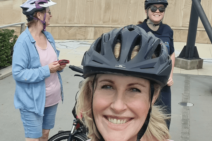 Kay McGrath smiles during the EcoTekkSC River to River e-bike tour at the HMAS Brisbane Memorial, Alexandra Headland, with other participants in the background making new friends.