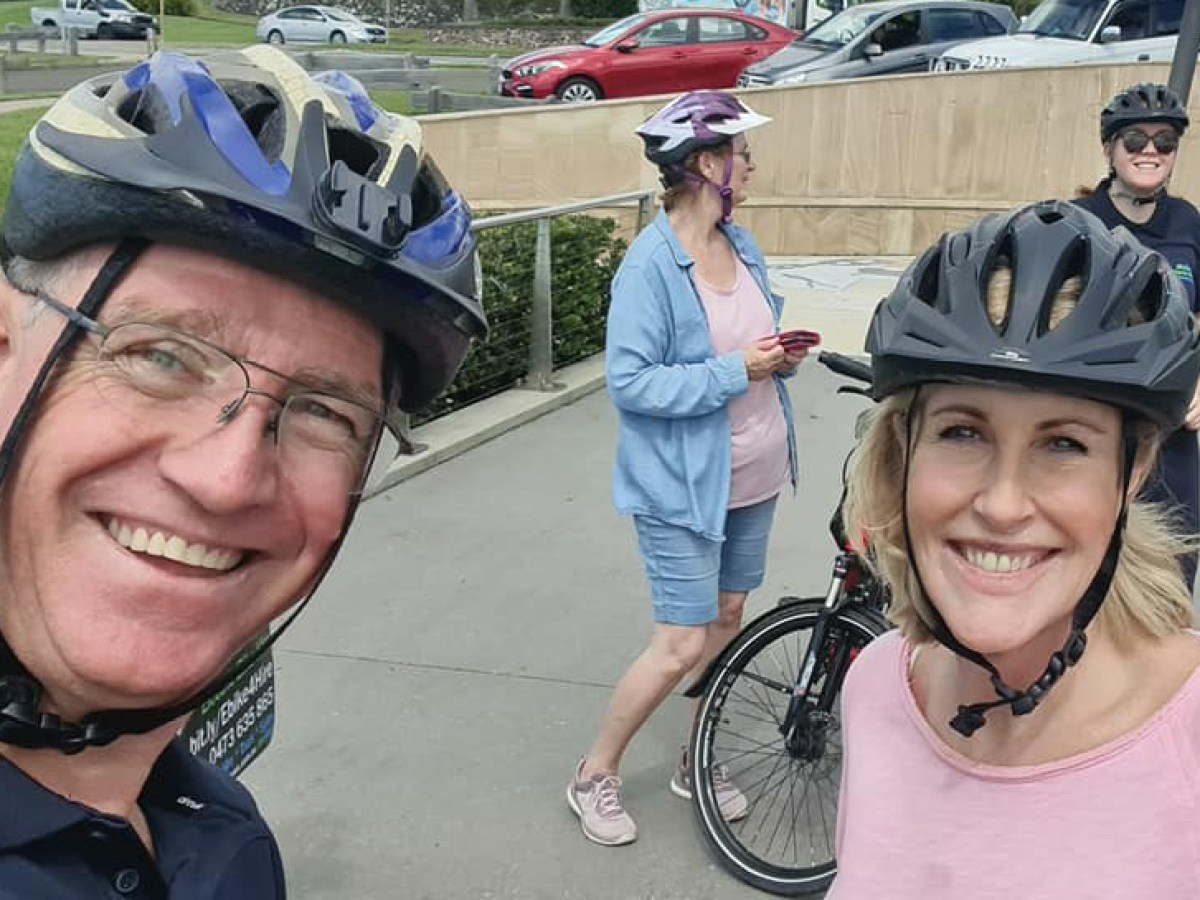 Stuart takes a selfie with Kay McGrath at Alexandra Headland during the River to River e-bike tour hosted by EcoTekkSC, with other participants smiling in the background at the HMAS Brisbane Memorial.