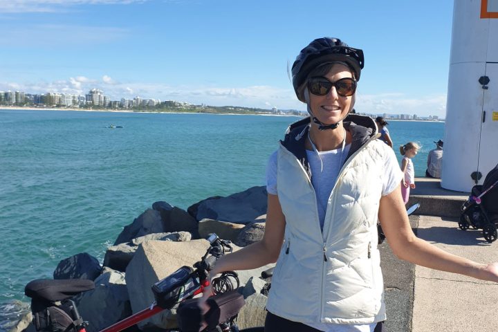 An e-bike rider beams with joy on the River to River Tour at the Mooloolaba Spit Rock Wall, overlooking the turquoise blue waters of Mooloolaba Bay and the Alex Heads skyline in the distance.