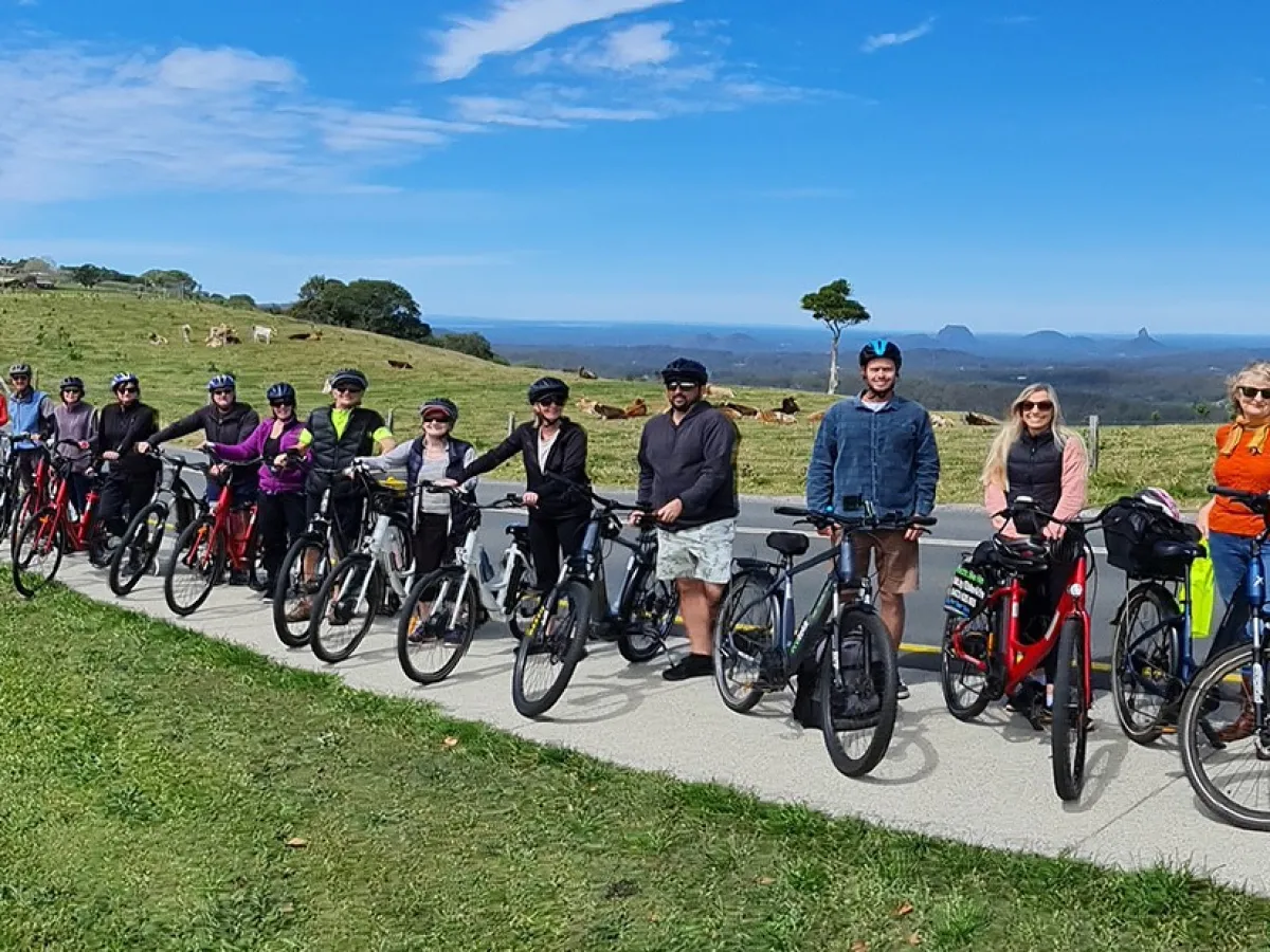 A group of e-Bike cyclists smiling and laughing in front of One Tree Hill on Mountain View Road in Maleny, with the Glasshouse Mountains visible in the background.