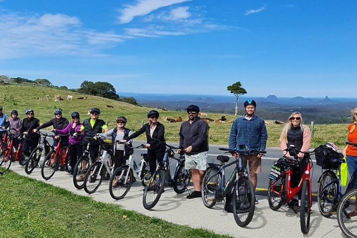 A group of 16 riders on EcoTekkSC e-bikes during the Maleny Magic Tour, stopping in front of the iconic One Tree Hill on Mountain View Road with the Glasshouse Mountains in the background.