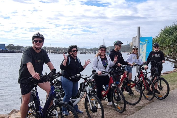 Six riders from Maple Weddings and Events with EcoTekk Sunshine Coast staff, smiling and waving in front of the coastal pathways sign at La Balsa Park, Buddina, during a bespoke e-bike tour.