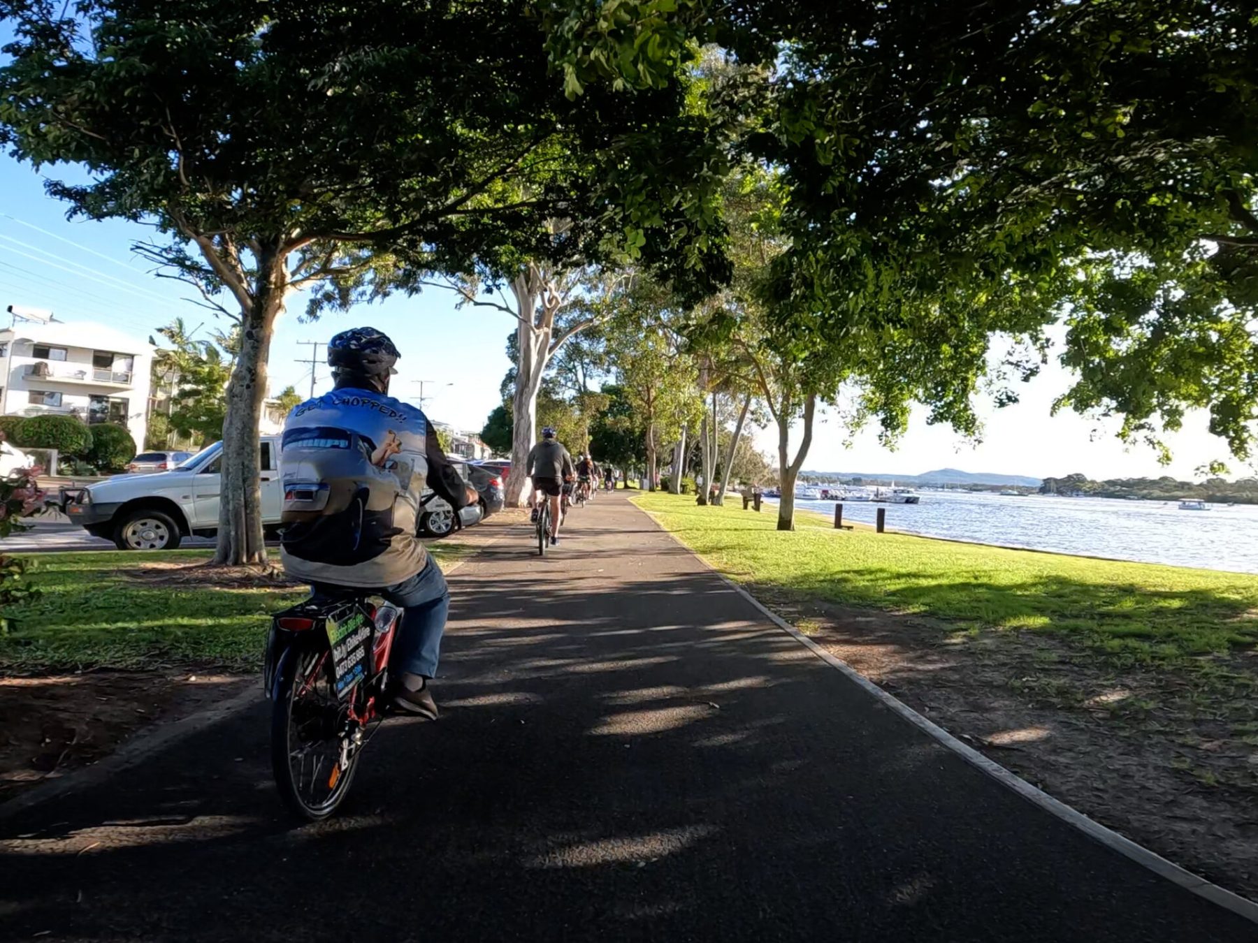 Riders on an EcoTekk e-Bike Tour along Gympie Terrace, Noosaville, with Pelican Beach Park and turquoise waters of the Noosa River under clear blue skies.