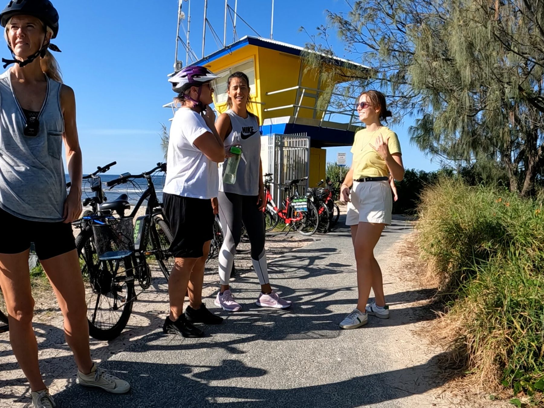 Corporate group of 20 riders on a bespoke EcoTekk Noosa e-bike tour stopping at Noosa River Groyne in Noosa Spit Recreation Reserve. Three female riders wait for a male colleague to remove his helmet before walking to the end for a group photo.