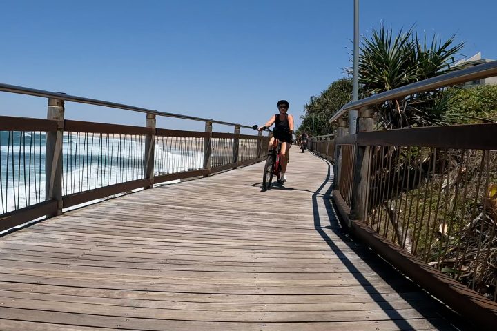 Husband and wife, with the wife leading, riding the coastal pathway on the ecoTekk Sunshine Coast Easy Peasy Rumba Golden Beach e-bike tour past Happy Valley Foreshore Reserve boardwalk, with Coral Sea waters and Bribie Island in the background.