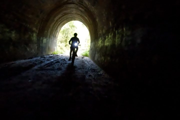 Silhouetted e-MTB rider entering the Dularcha Railway Tunnel, with the northern arch entrance illuminated by background light.