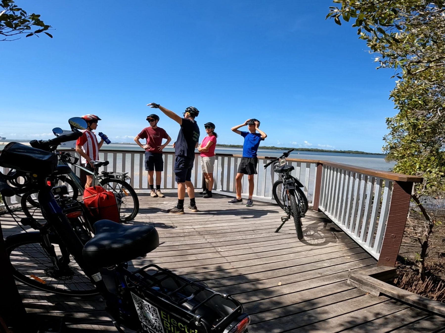 view of a family of four, Mum, Dad, and two teenage boys, taking a break at Monash Park Mangrove Boardwalk during the ecoTekk Sunshine Coast Golden Beach e-bike tour, with turquoise waters of Pumicestone Passage and a marine biologist guide discussing coastal ecosystems.